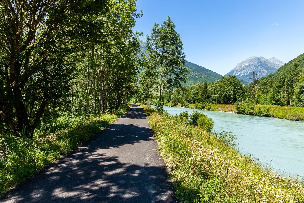 Voie cyclable avec ombres, bordées arbres, près d'une rivière et vue sur les montagnes